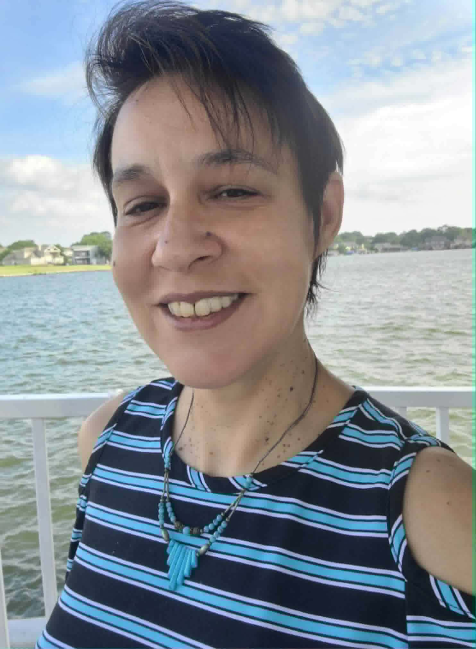 Person smiling by a lake with striped top and turquoise necklace.